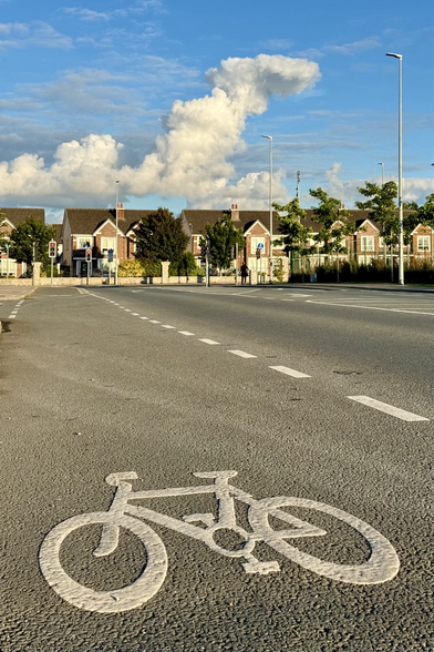 A low-down shot with the painted bike logo on a cycle lane in the foreground, some semi-detached brick homes in the mid-ground and a rich blue sky with big puffy white clouds above