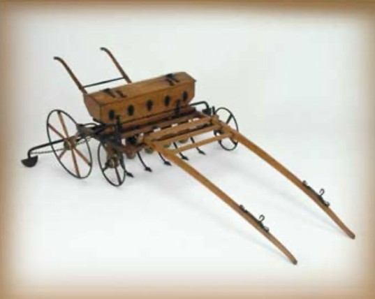 A vintage wooden horse-drawn seeder with metal wheels and two long handles.