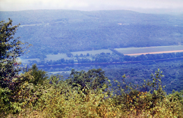 A hazy whitewashed summer sky. We are high atop a ridge crest looking out over treetops down a few hundred feet to a river valley below. Most it is wooded, the rest is pastures and fields. Long, flattish ridges rise above the far side of the valley as the landscape gradually dissolves into the haze and humidity.