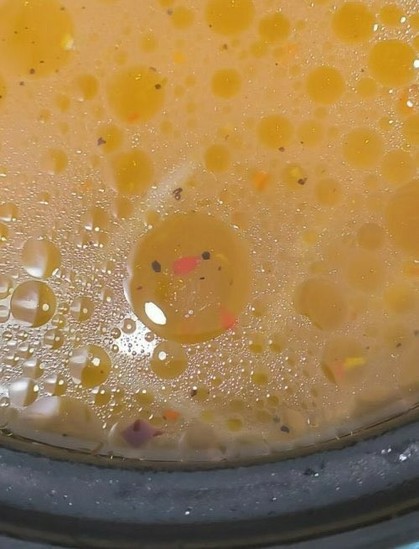 A bowl of clear yellow soup with bright yellow fat droplets on the surface. One of the droplets seems to have two eyes (specks of pepper) and an orange nose and feet (specks of carrot). It looks like a baby chicken (chick)