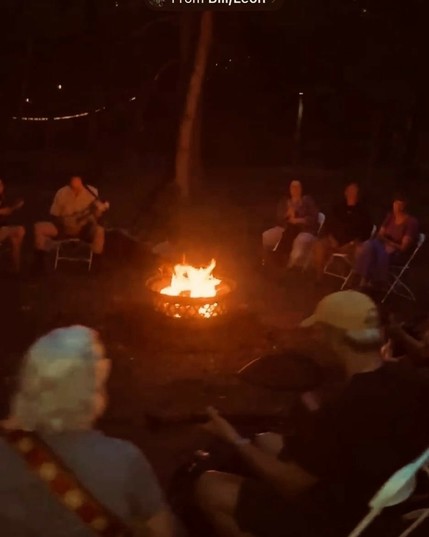 A group of people sitting around a campfire at night, playing guitars.
