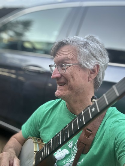 A person with gray hair and glasses smiling while playing a banjo, wearing a green t-shirt and sitting in front of a parked car.
