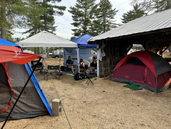 Camping area with several tents, people sitting under canopy shelters, and a wooded background.