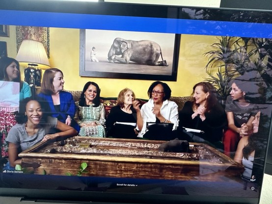 Gloria Steinem in her living room sitting on a large couch surrounded by 8 other women of various races and ages. They seem happy and are laughing at something one of them just said.