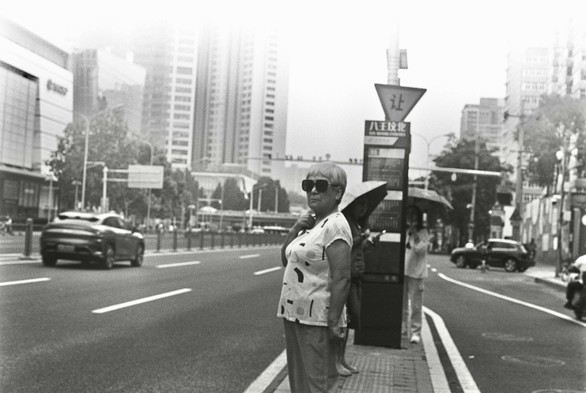 A black and white image of a person waiting for a bus at a bus stop while a car driving by.