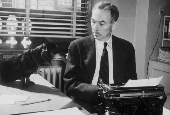 Black & white photo of E. B. White at his typewriter in his office at The New Yorker. He is looking at his dachshund, Minnie, who is sitting to his right on his L-shaped desk and looking back at him. (Photo credit: New York Times Co./Getty Images)