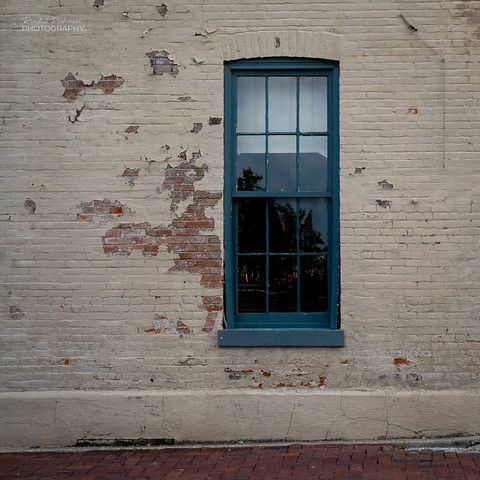 A tall green window is set in a brick building with peeling tan paint.