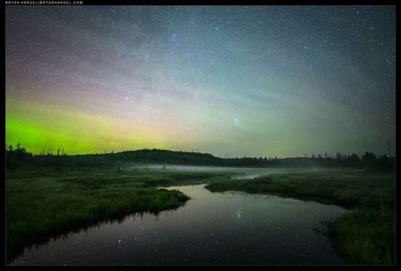 A creek reflecting stars leads out to a star sky filled with northern lights.