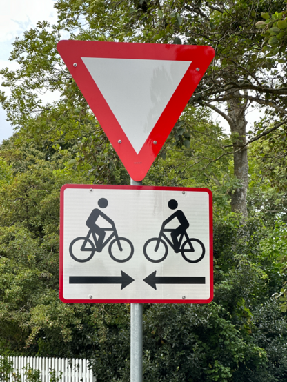 A “give priority sign” (inverted white triangle with red edge) with below a sign depicting two bicycles with riders in opposite directions and two arrows below the bicycles pointing to each other. Bicycles and arrows are black on a white rectangle sign with a thin red edge. 