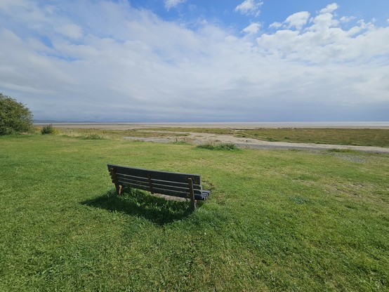 A bench on a swathe of grass looking out over a flat landscape of sand and water.