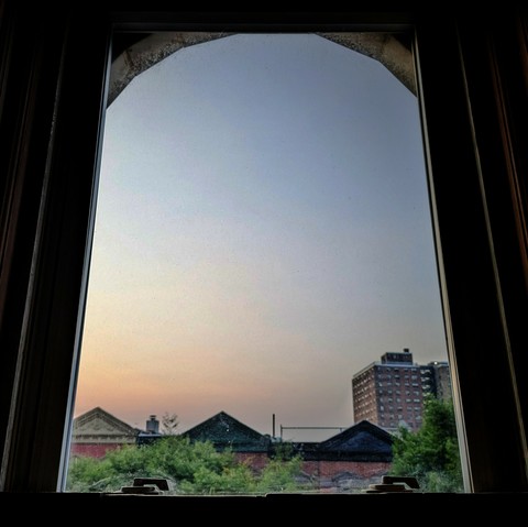 Looking through an arched window twenty minutes after sunrise the cloudless sky fades diagonally from peach on the bottom left to slate blue on the upper right. Pointed roofs of Harlem brownstones with red brickwork are across the street, and a taller apartment building can be seen in the distance. The green tops of two trees are filling the frame on the bottom and right.