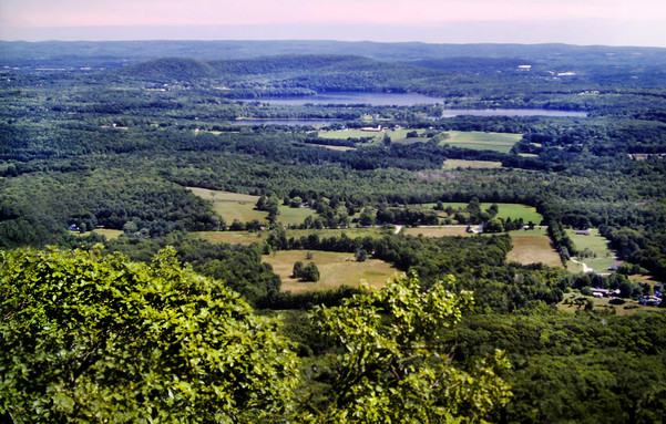 We are standing on the edge of a plateau looking over treetops down into a sun-drenched valley hundreds of feet below. The valley floor is relatively flat, although wooded hills rise in the distance along the horizon. Most of the valley floor is wooded, too, but there are green fields, rural lanes, some houses and a large pond visible. The sky is bright and soft blue with gauzy, hazy clouds.