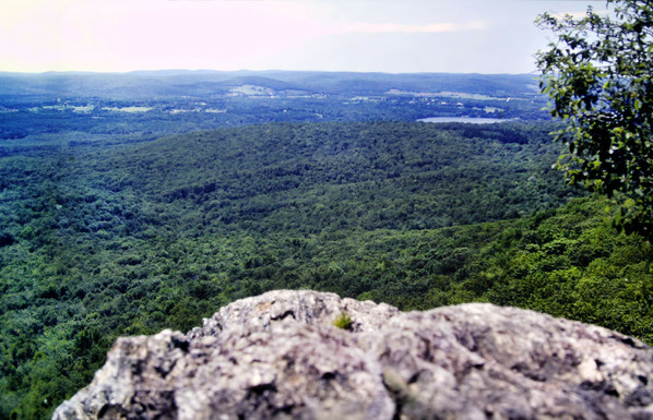 We are standing atop cliffs of reddish gray rock looking down into a sun-drenched valley hundreds of feet below. The valley floor is relatively flat, although wooded hills rise in the distance along the horizon. Most of the valley floor is wooded, too, but a few miles distant there are a few green fields, some houses and a small pond visible. The sky is bright and soft blue with gauzy, hazy clouds.