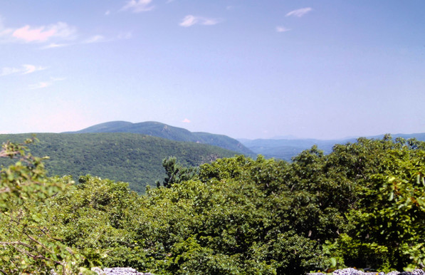 Beneath a soft blue sky with a few clouds, we are atop a mountain looking over treetops northward along the ridge ahead as it passes over a series of mostly wooded rounded peaks. We can see for miles in the clear, dry air. Lines of hills and other mountains run across the horizon.