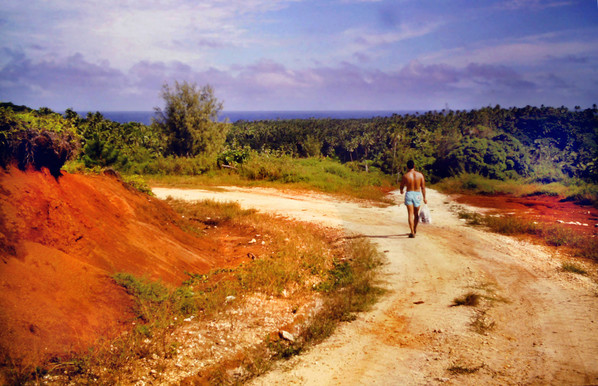 A man in a bathing suit carrying a bag is walking down a light colored dirt road past a slope of bright red clay on the left. The road curves left and a smaller road leads straight ahead through a forest of palms and other tropical trees towards the distant blue ocean visible along the horizon. The sky is a soft blue, with streaky white clouds and thicker clouds along the horizon.