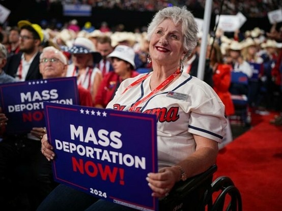 White middle-aged woman in a wheelchair holding a “Mass Deportation Now!” Trump sign at the Milwaukee RNC in July. 