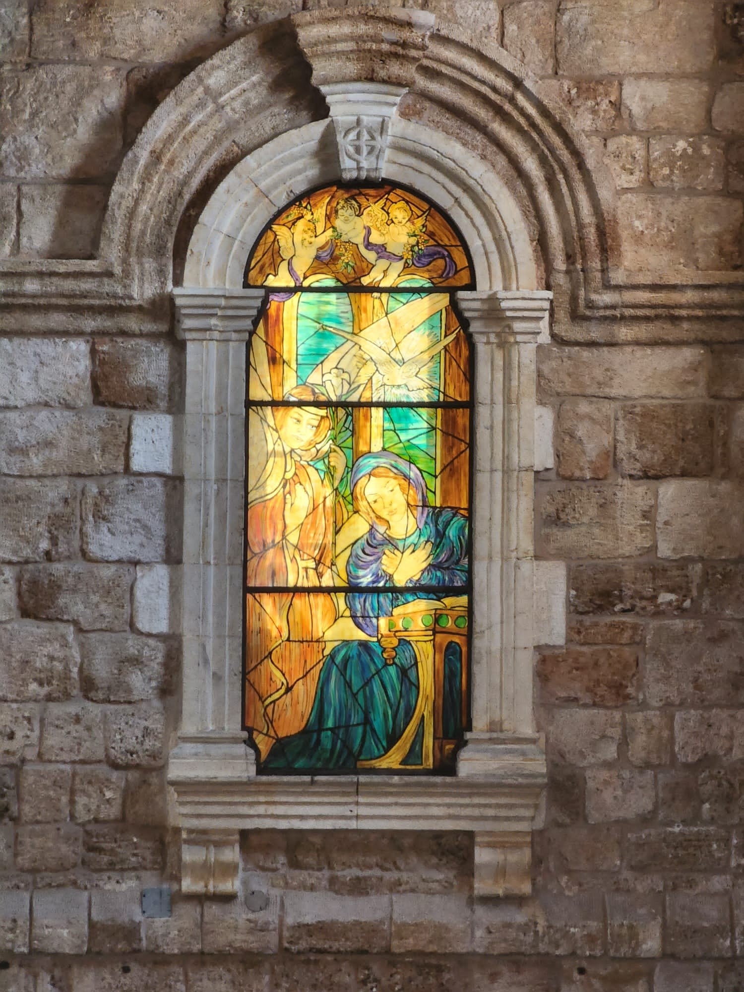 arched stained glass window about a meter tall and .5 meter wide. in mostly bright yellow and orange tints it depicts the Annunciation.