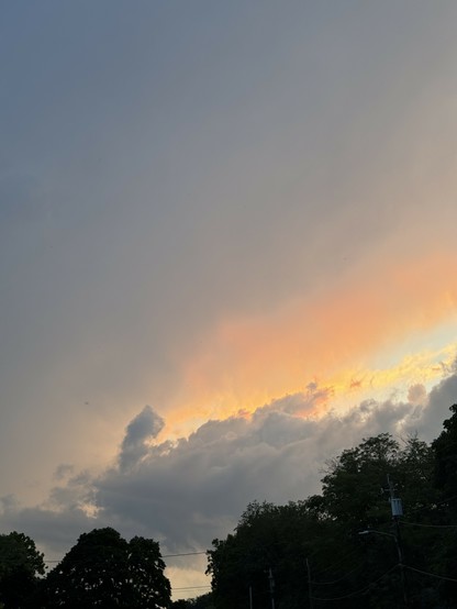 A sunset scene with orange hues peeking through a cloudy sky, silhouetted trees in the foreground, and power lines visible.