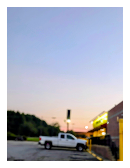 out of focus side view from the far end of a waffle house parking lot of a restaurant with white truck parked in front. the parking area is otherwise empty. in the background, store's sign, a grassy area, a treeline. the sky is pale orange at the horizon, pale gray/blue above.