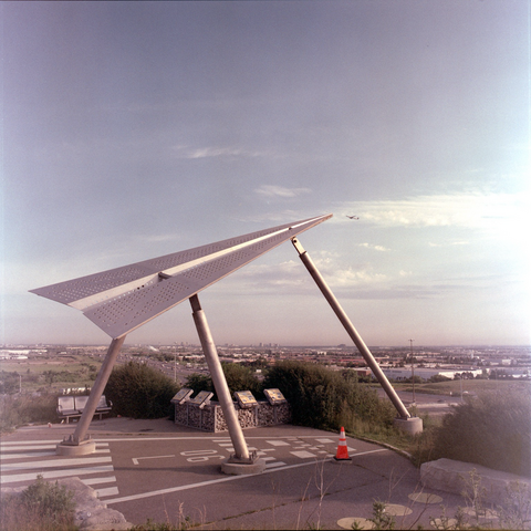 A metal sculpture depicts a giant paper airplane taking off from a hilltop in a landscape of mega highways and industrial parks. An actual airliner appears to approach the tip of the paper airplane as it rises into the morning sky.