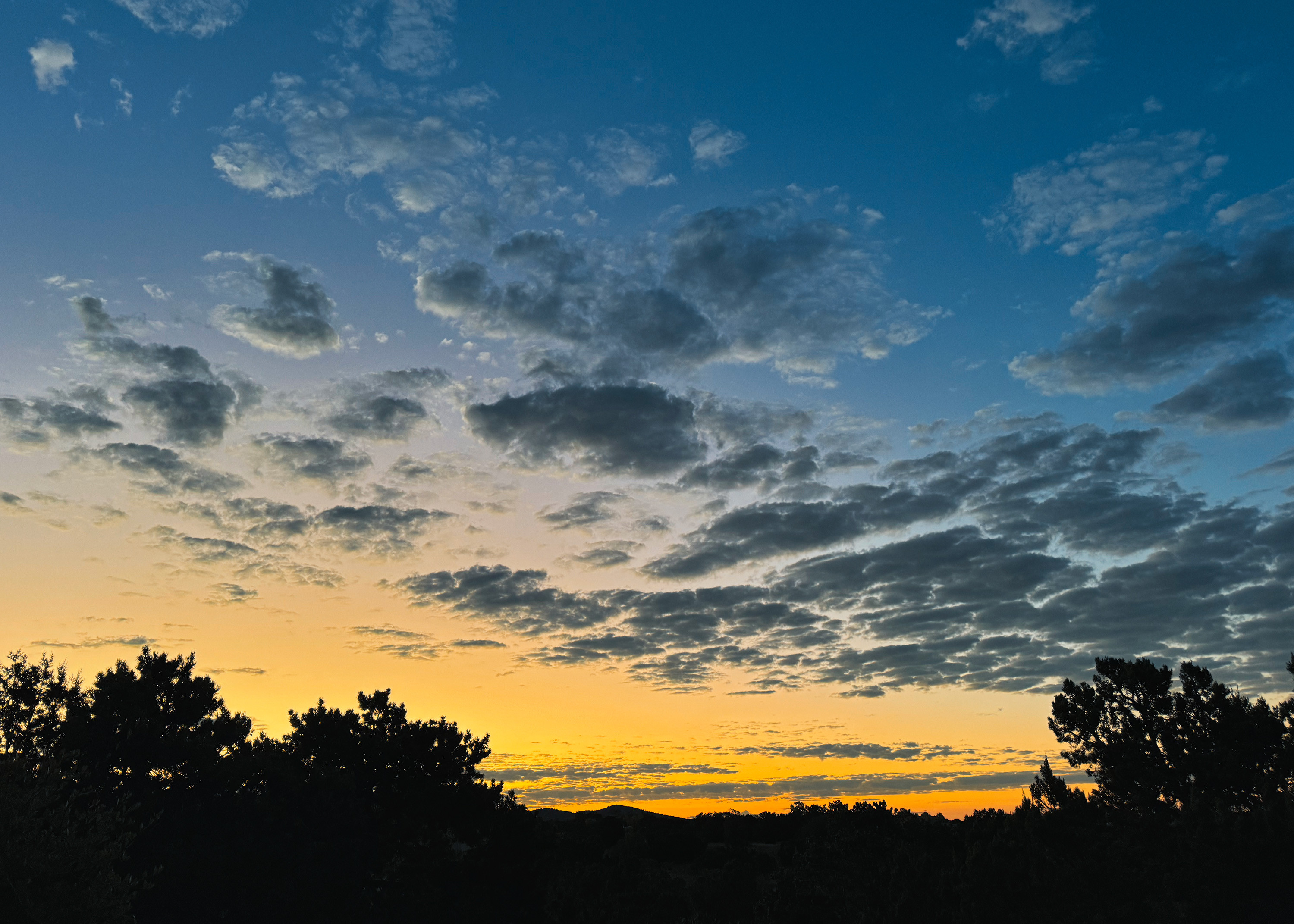 Dawn in Santa Fe, New Mexico. Orange sky at the horizon, blending to yellow, to dark blue overhead. Many small shreds of clouds across the frame, dark landscape shadows below.