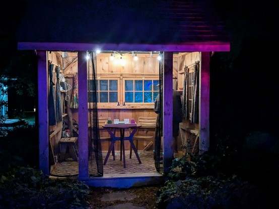 A small wooden building is lit up at night.
The building has a table set for a meal.
The lights are multi-colored with the building illuminated in purple.