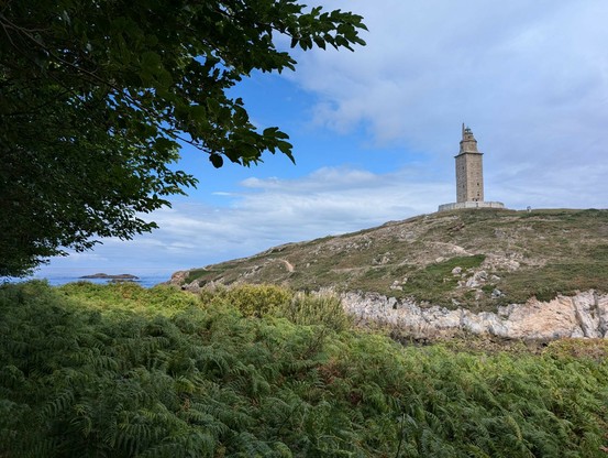 Landscape view of grass before a drop off to a bay out of sight, then cliffs and grassy lane above to where a tall tower stands. It's stone and square-based and without decoration, but has the look and feel of an old lighthouse. Trees overhang from the left of view. It's a bright day with blue sky and white clouds.