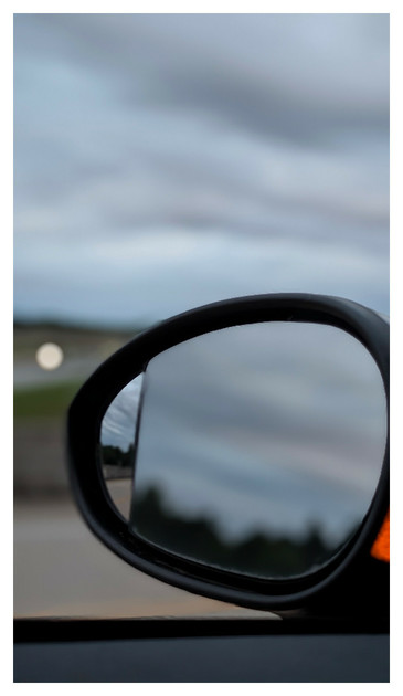 cloudy morning on a freeway overpass. though my driver's side window. close-up of rearview mirror gives 2 views of the area behind the car, colorful clouds, a cement road and treeline, the background is out of focus road, freeway median and a single white dot on the road in the distance.