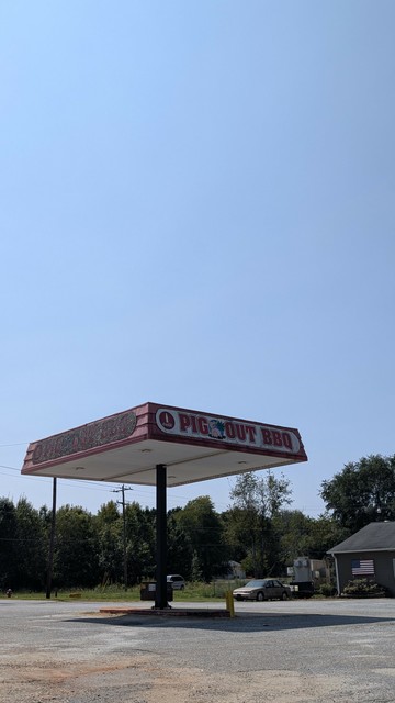 daytime. fuel station canopy with Big out BBQ on two sides. in the mid distance. a car with barbecue smoker next to a building with an American flag out front trees around, etc. if anyone can provide an alt text for this. it would be much appreciated.