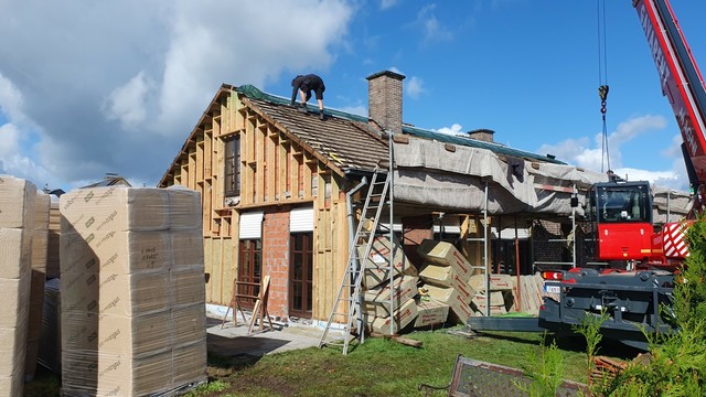 A detached house with the facade and the roof shingles removed. In front of the house are stacks of insulation material, scaffolding and a crane. On the dismantled roog a construction worker is attaching a green canvas.