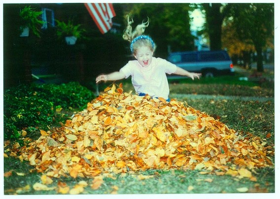 Color image of a young girl leaping into a leafy pile. Image shows a conical pile of leaves in a yard, a delighted girl leaping into them, eyes closed, smiling and laughing, blonde hair floating behind her.