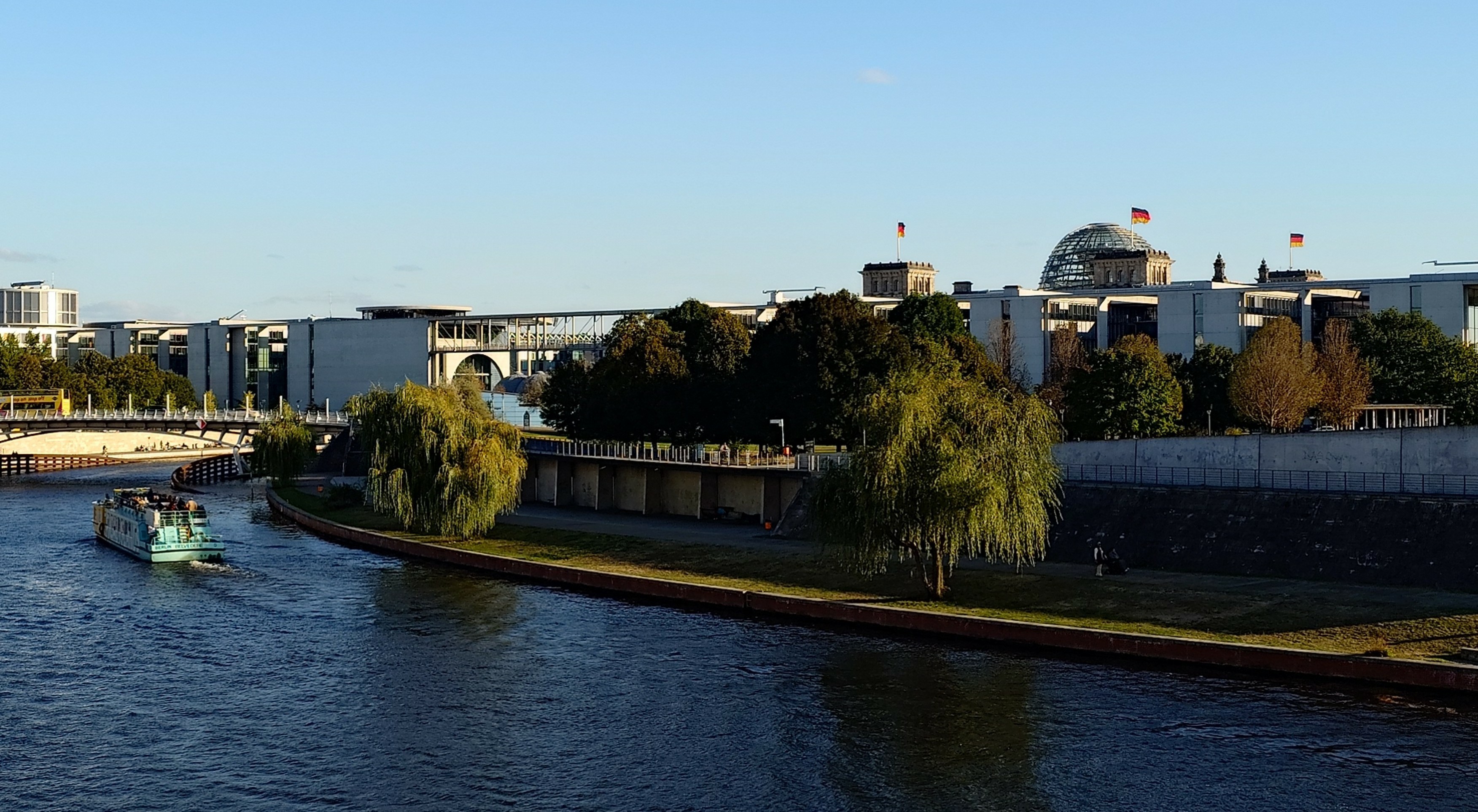 Behind the river Spree in downtown Berlin, a tourist boat cruising along and spanned by a bridge, some trees and lawn on its shores, in the background the building of the German parliament with its central glass cupola and German flags flying, under a <br />cloudless sky.