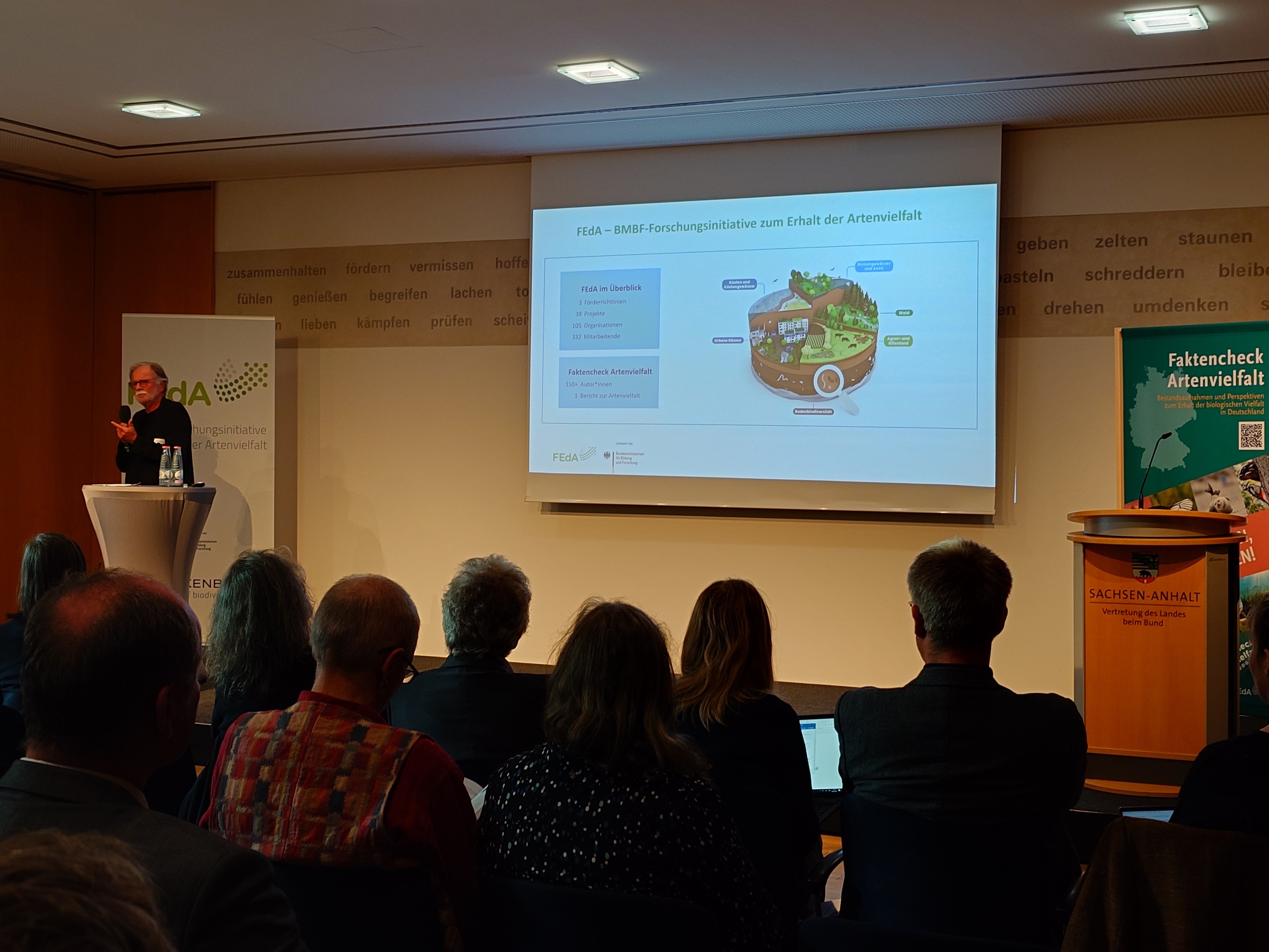 A crowd is listening to Volker Moosbrugger, German biodiversity scientist, introducing the Fact Check Biodiversity Germany. He is walking behind a podium and showing an overview slide.