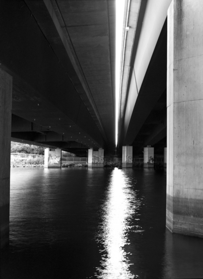 We are standing under two bridges over a smooth river, barely separated by a crack of overexposed sky. The light paints a bright divide across the dark water, as if the stream were being torn apart. 