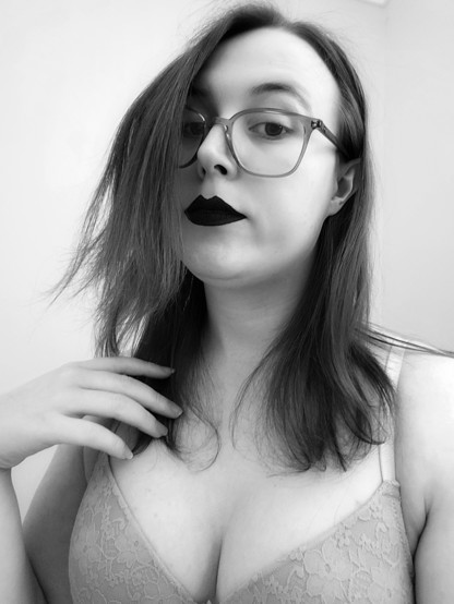 Black & white selfie of Revy
Her hair is fluffy and luscious 
She has black lipstick and is wearing glasses
Her hand rests softly on her shoulder that holds some of her hair.
She's wearing a bra showing cleavage