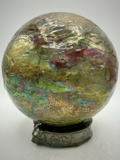 A sphere on a base with a blend of iridescent hues, golden tones, deep reds and shimmering greens.