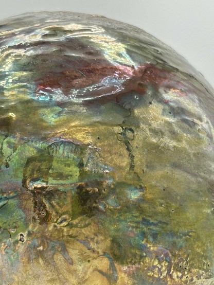 A close up of a sphere with a blend of iridescent hues, golden tones, deep reds and shimmering greens.