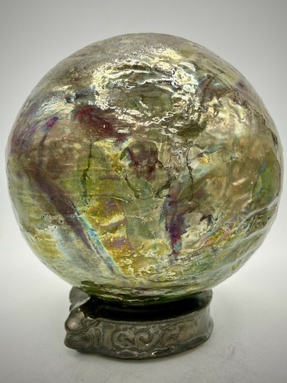 A sphere on a base with a blend of iridescent hues, golden tones, deep reds and shimmering greens.