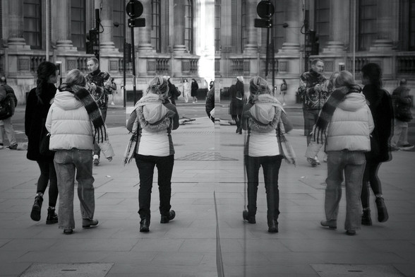 A high contrast black and white photo of three women walking away from the camera and a couple walking towards the camera, all reflected in a window at the right hand side of the camera.