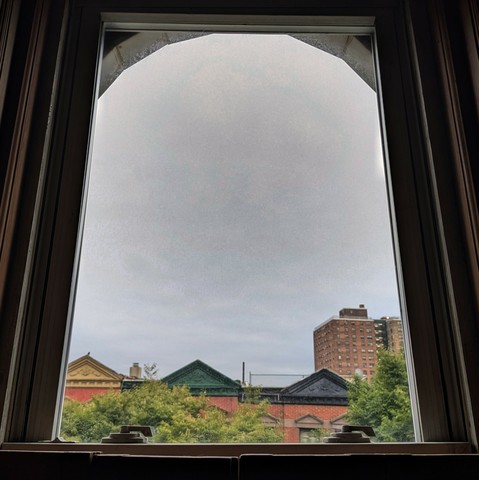 Looking through an arched window at the moment of sunrise the sky is full of grey clouds. Pointed roofs of Harlem brownstones are across the street, and a taller apartment building can be seen in the distance. The green tops of two trees are entering the frame on the bottom and right.