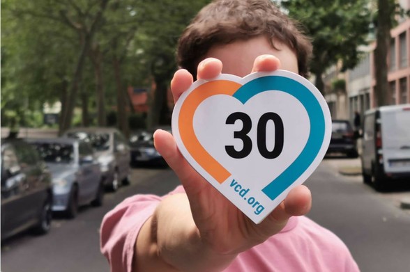Auf dem Bild ist ein junger Mann zu sehen, der auf einer Straße steht, und einen herzförmigen Tempo-30-Aufkleber im VCD-Design in die Kamera hält.