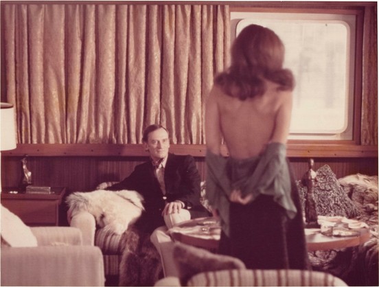 A woman with a bare back stands facing a seated man in a vintage-decor room.