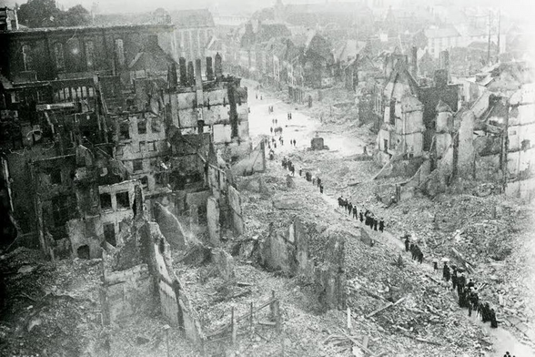 A black and white photograph showing a bird's eye view of a square surrounded by ruins.
A line of people is walking through the rubble towards the square.