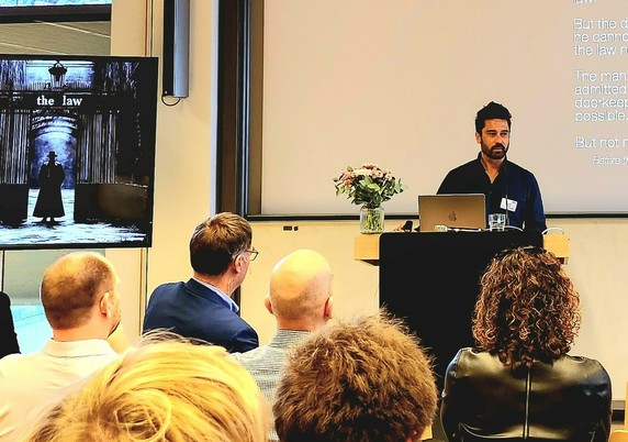 The picture shows Casey Mock giving a lecture. You can see an attentive audience with their head turned towards the speaker. To his left you can see a picture of a person standing in a judicial hall with "the law" written above.

