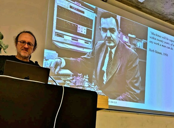 The picture shows Bernhard Schölkopf giving a lecture. Behind the speaker you can see a picture of writer Luis Borges projected onto a screen.