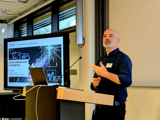 The picture shows Mike Schäfer giving a lecture. To his left, you can see a screen showing the first slide of his presentation. The title reads "Science Communication in the Age of AI".