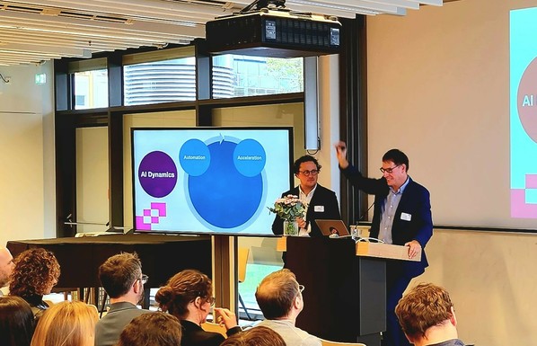 The picture shows Markus Gottschling and Olaf Kramer standing behind a podium and giving a lecture. A screen to their left shows a snippet from their presentation reading "AI Dynamics", "Automation" and "Acceleration".