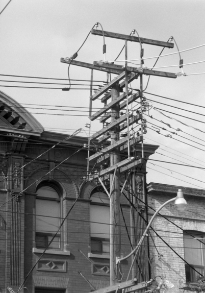 A very busy utility pole, with multiple cross pieces (even the cross pieces have crosspieces), rises in front of an important looking brick building of the early 1900’s. 