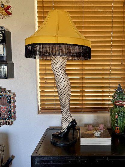 iPhone photo of our yellow Leg Lamp with black fishnets sitting on a dark metal console in front of a window with wooden slatted shades, there’s folk art & a pay phone hanging on the wall to the left & books & a handmade green character jug sitting on the console to the right