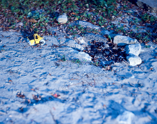 A yellow plastic toy car, stylized like a construction vehicle, has been forgotten behind and left in the sand near an extinguished beach fire pit. 